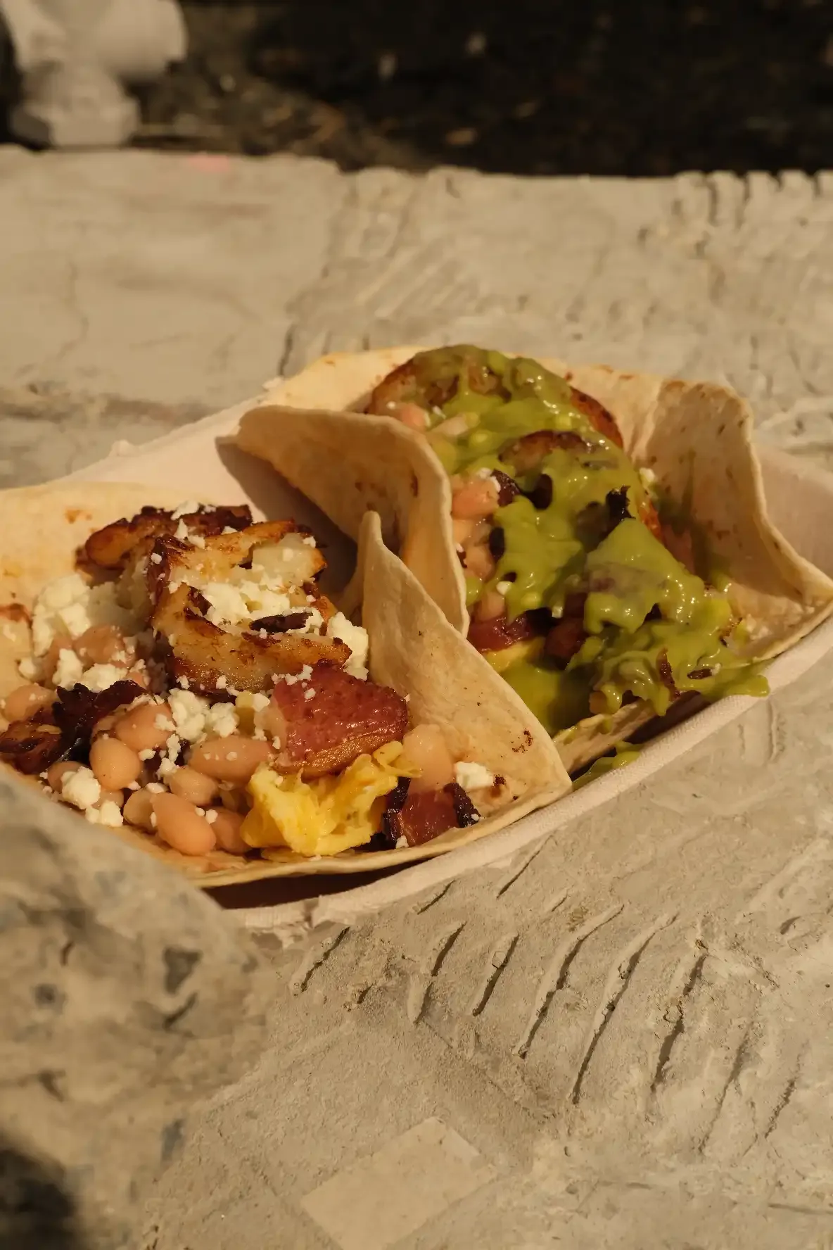 Two breakfast tacos in a paper boat with scrambled egg, white beans, bacon, crispy potato and queso fresco