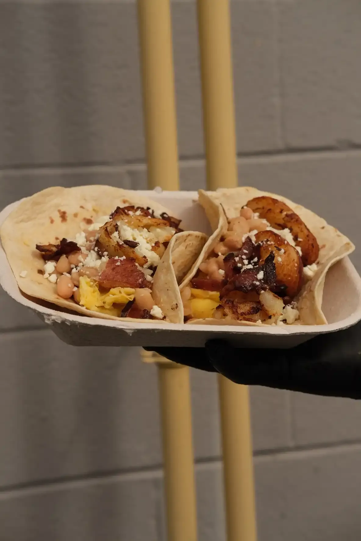 A gloved hand holding two handheld breakfast tacos in a paper boat, filled with scrambled egg, white beans, bacon, crispy potato and queso fresco