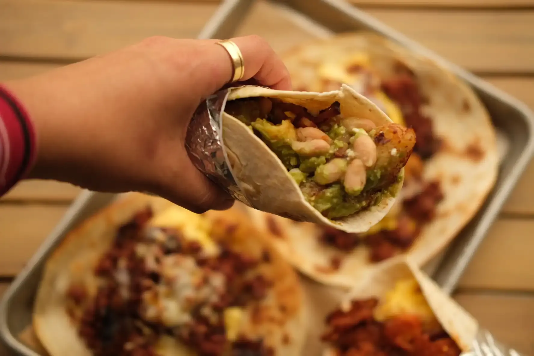 A hand holding a foil-wrapped breakfast taco filled with scrambled egg, white beans, crispy potato and green salsa