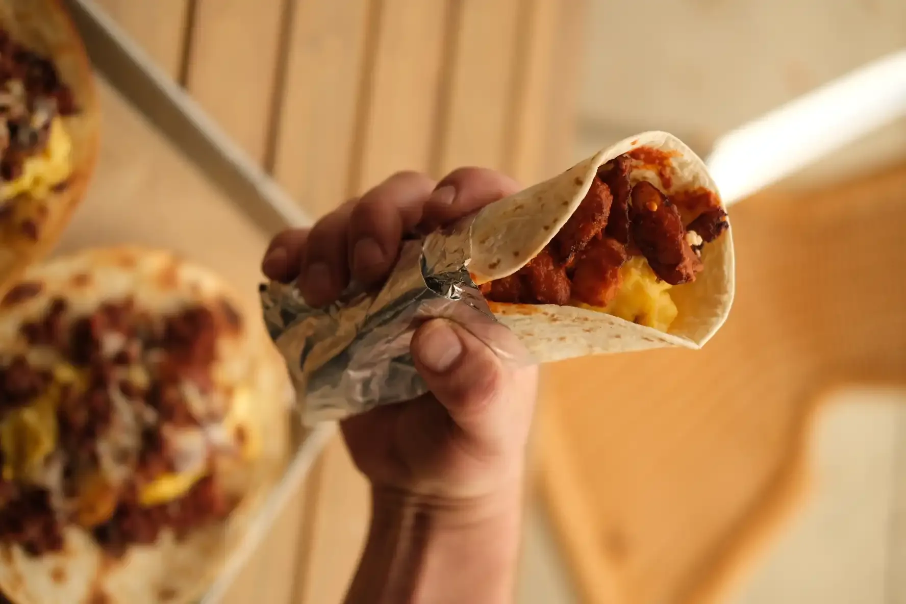 A hand holding a foil-wrapped breakfast taco filled with red salsa, scrambled egg and crispy potato, seen from below