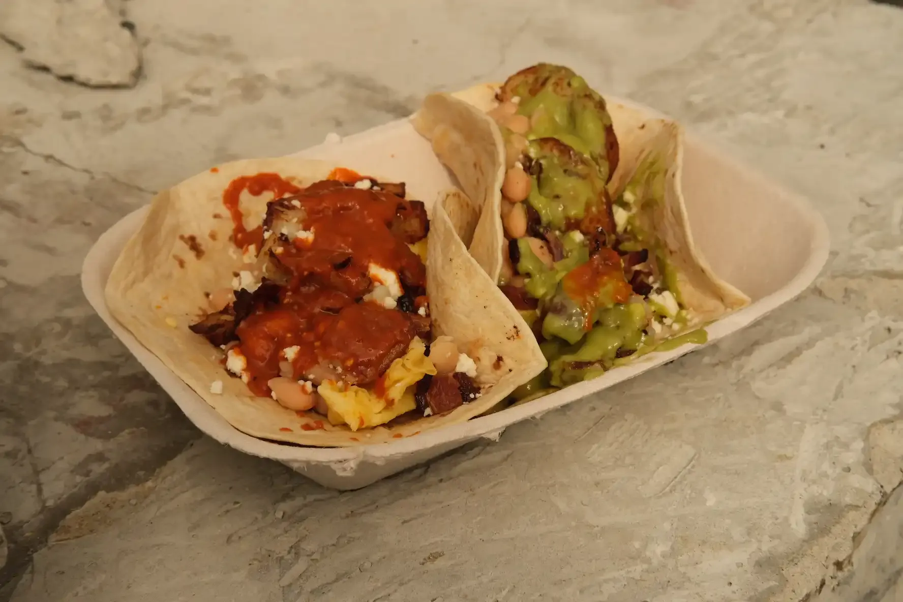 Two breakfast tacos in a paper boat on a stone surface, one topped with salsa roja and one with green salsa