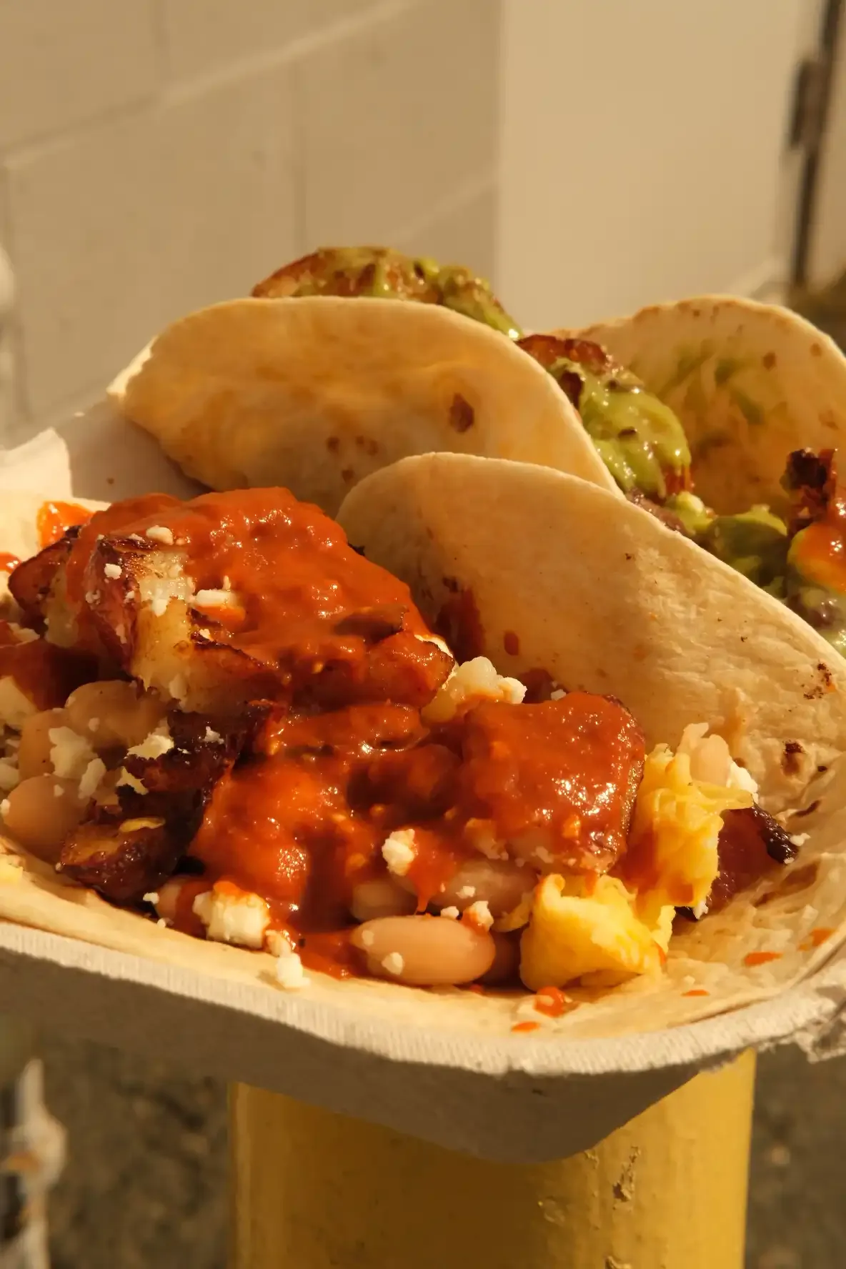 Close-up of a breakfast taco loaded with crispy potato, white beans, scrambled egg and queso fresco under salsa roja, beside a taco with salsa verde