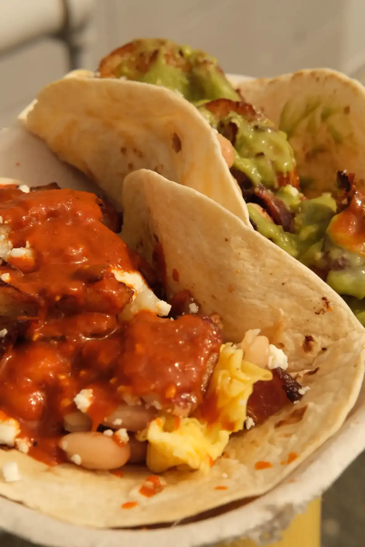 Breakfast tacos with scrambled egg, white beans, crispy potato and queso fresco under salsa roja, beside a taco topped with salsa verde
