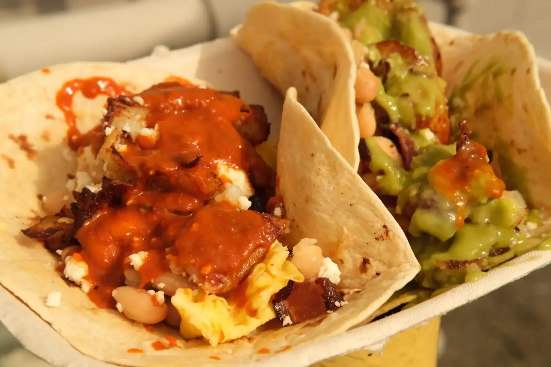 Close-up of a breakfast taco with scrambled egg, white beans and queso fresco under salsa roja, beside a taco topped with salsa verde