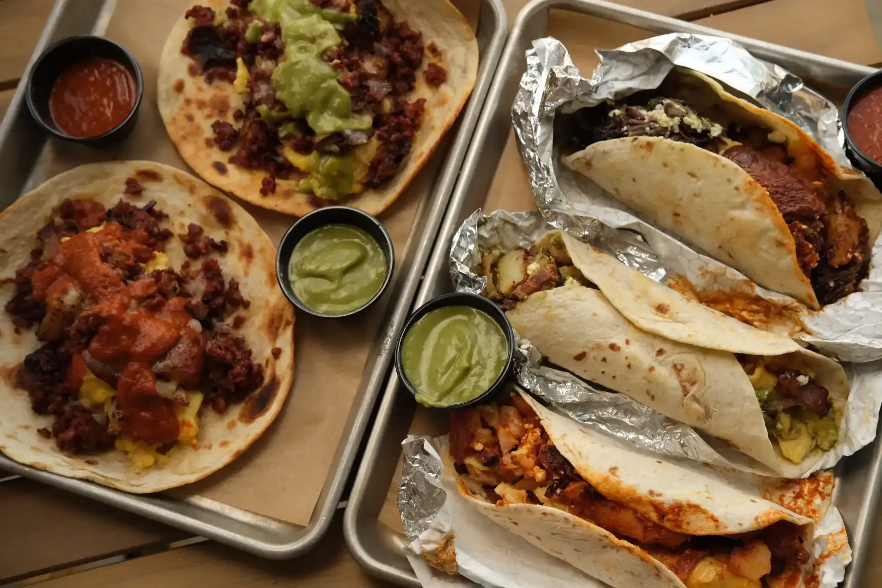 Two metal trays of chorizo and egg tacos with cups of salsa roja and salsa verde, beside three foil-wrapped breakfast tacos