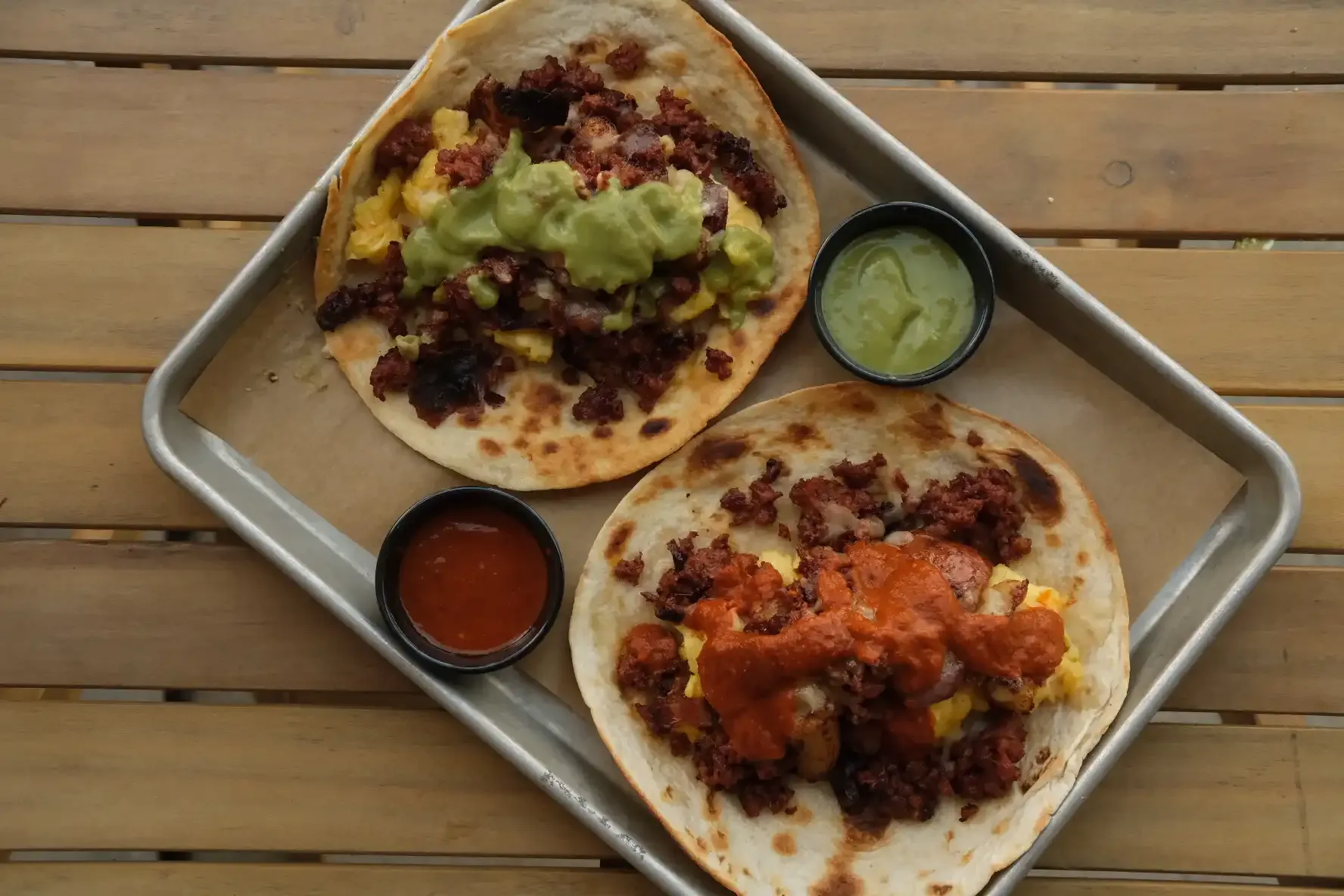 Two chorizo and egg tacos on a metal tray with cups of salsa verde and salsa roja on a wooden table